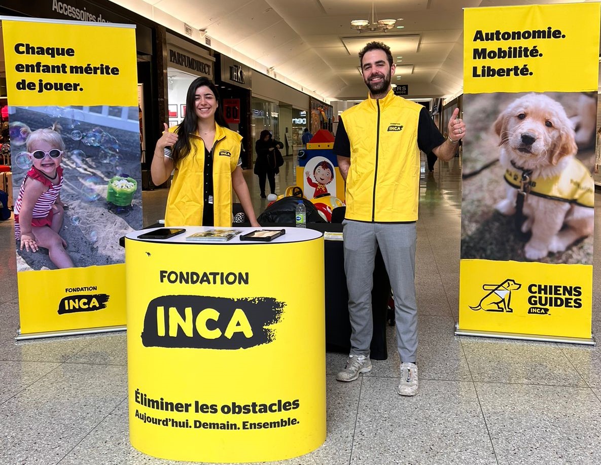 Deux solliciteurs debout devant un présentoir d'INCA dans un centre commercial.