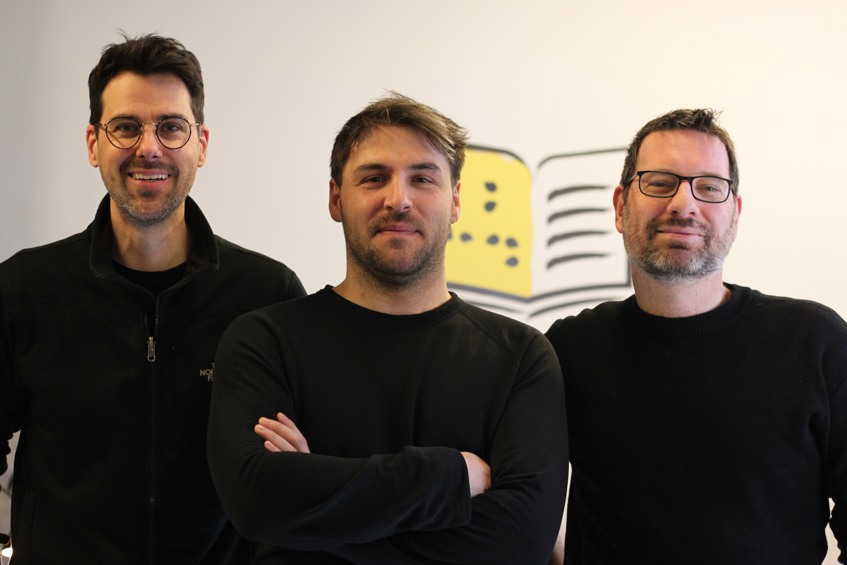 Charles, Simon et Emmanuel sourient devant l'icône d'un livres accessible aux couleurs d'INCA.  Ils portent tous les trois un chandail noir.