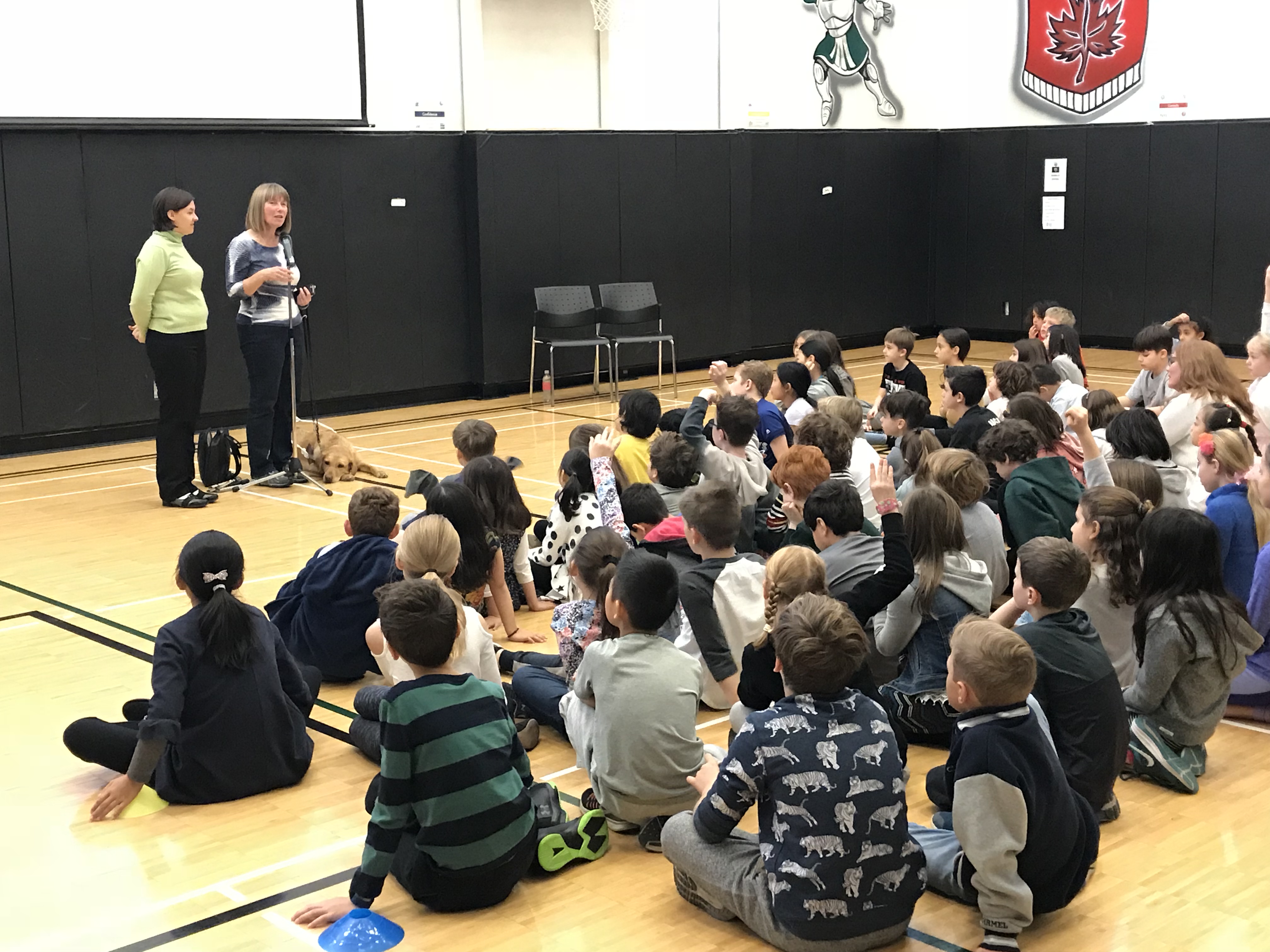 Two young women and a guide dog stand at the front of a gymnasium and give a presentation a group of kids seated on the gym floor. 