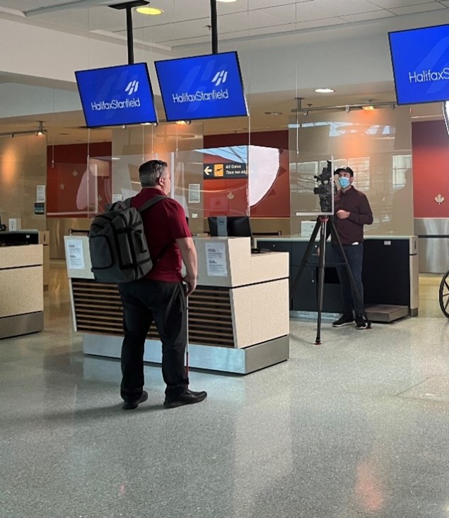 Duane Morgan, CNIB’s Vice President of Atlantic Canada, stands in front of an airport kiosk with a cameraman filming in the background.