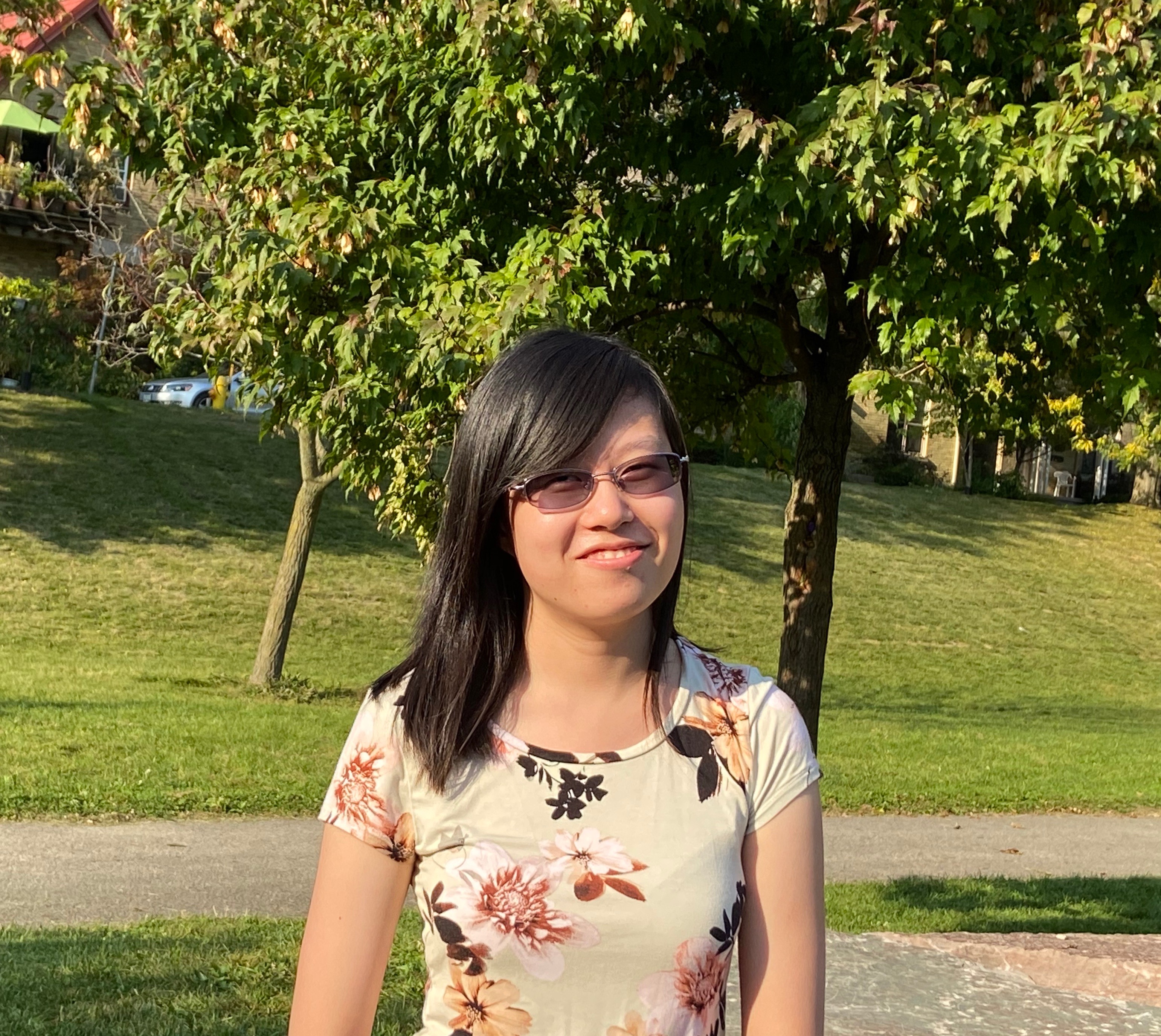 Lily is outside, standing in front of two lush green trees. She is smiling and wears a floral top and sunglasses. 