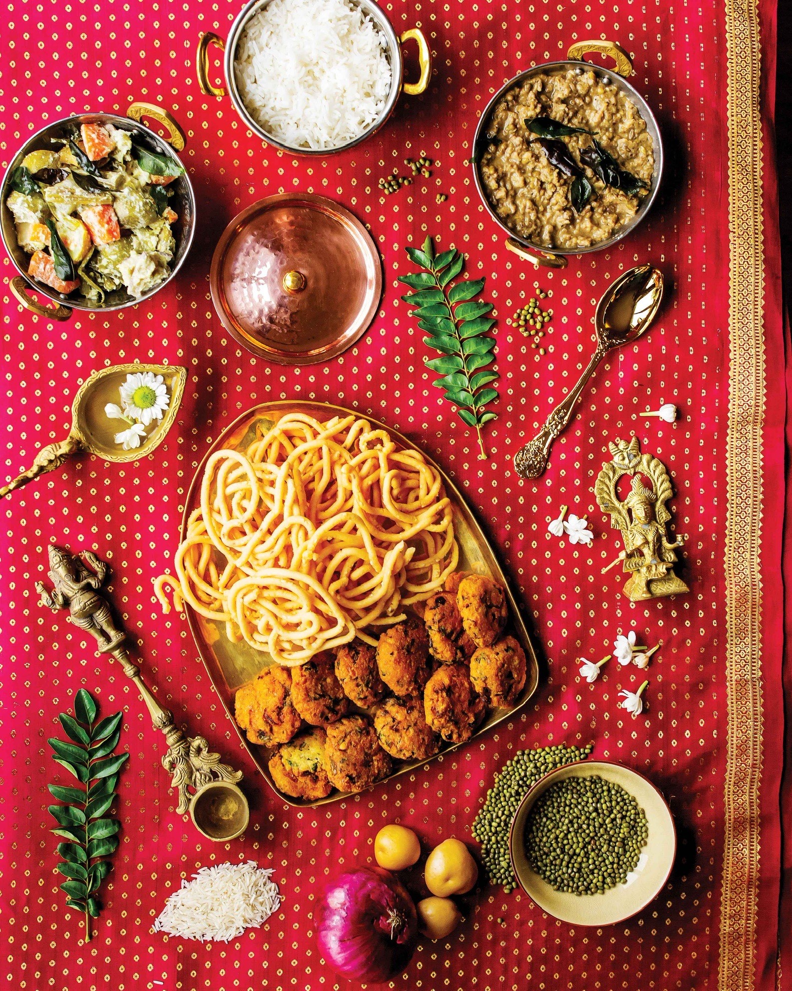 Several dishes in the book including murukku (crispy snack), vada (a savoury fried snack), avial (coconut curry with vegetables), basmati rice, and dhal (lentils) appear in gold dishes on a magenta and gold silk saree. Colourful ingredients, jasmine flowers, and a gold statue are found on the table setting amongst the dishes.