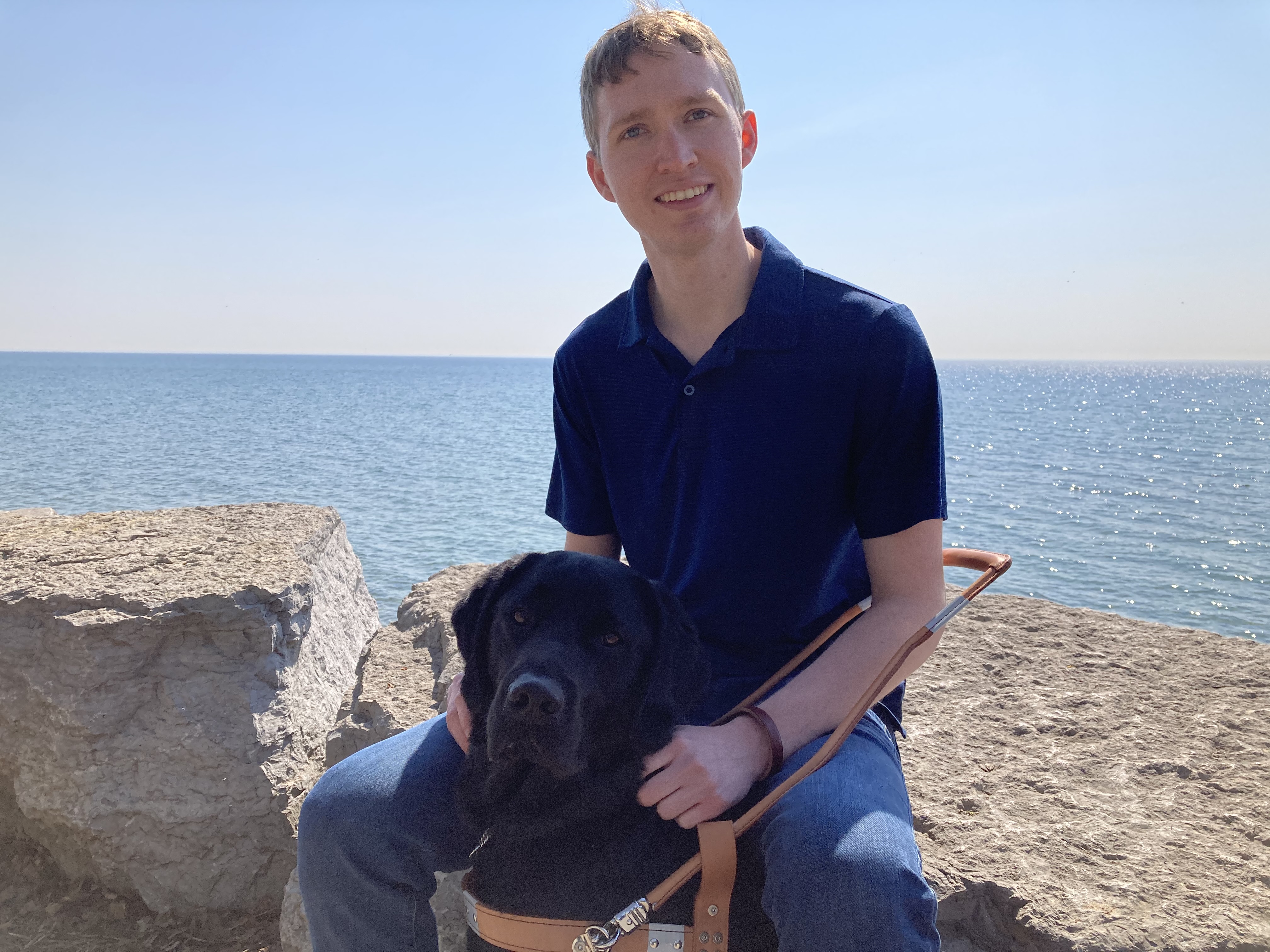 Jack McCormick and his guide dog, Baloo, a black Labrador retriever, sitting on large rocks in front of the ocean and smiling for the camera