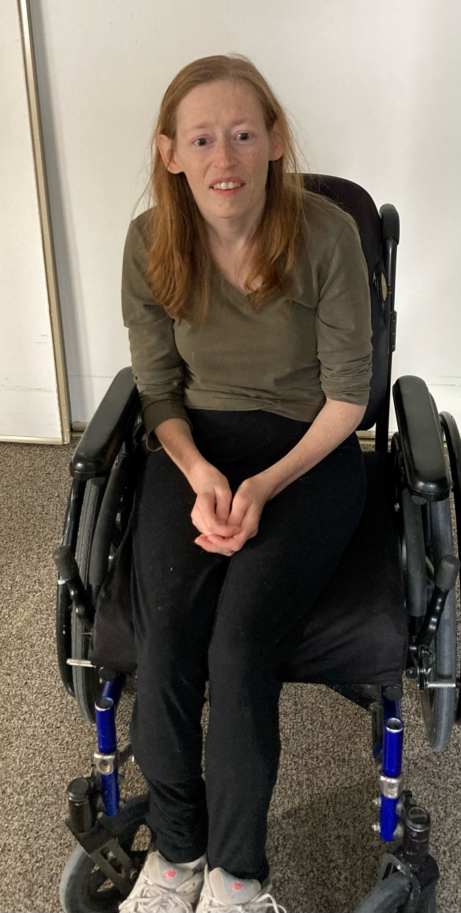 Julia sourit et pose pour une photo. Elle est assise dans un fauteuil roulant et porte une chemise verte et un pantalon noir.