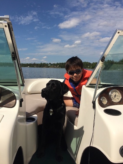 Keaton est assis sur un bateau dans l'eau avec son bras autour d'Indy, un labrador golden-retriever noir, qui est assis sur le bateau à côté de lui.