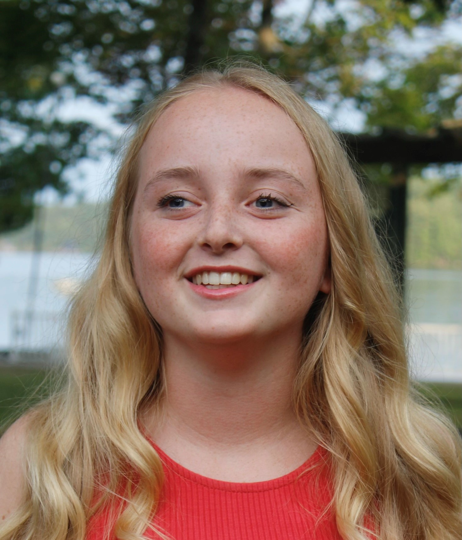 Portrait de Maggie, souriante, pris en extérieur à Lake Joe, près du front de mer. Maggie est une jeune femme aux longs cheveux blonds qui porte une chemise orange. 