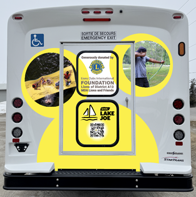 ​​​​La porte arrière du nouveau minibus. La porte comporte un décalque lumineux avec des motifs abstraits de bulles jaunes et des photos de campeurs participant au tir à l'arc et profitant du lac. Au centre de l'autocollant figure le texte suivant : « Generously donated by Lions Clubs International Foundation, Lions of District A12, MDA Lions and Friends », accompagné du logo du Fonds du Lions Club International, du logo Camp Lake Joe d’INCA et d'un code QR.