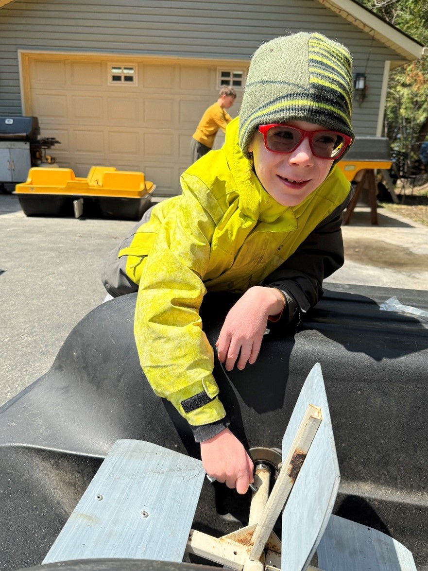 Evan s'appuie sur le bateau à aubes dans l'allée de leur maison. Il porte une veste jaune, un chapeau vert et des lunettes rouges. Son frère Noah se tient à l'arrière-plan, travaillant sur un autre bateau à aubes.