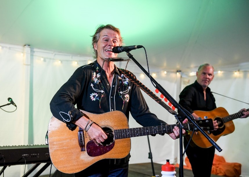 Jim Cuddy performing live at CNIB Lake Joe. He is on stage, playing a guitar and singing into the microphone. Another member of his band plays guitar in the background.