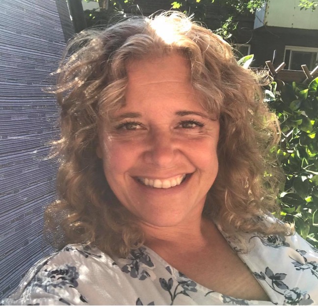 Un égoportrait de Laura Mason.  Elle est assise à l’extérieur, au soleil, avec une plante verte luxuriante derrière elle. 