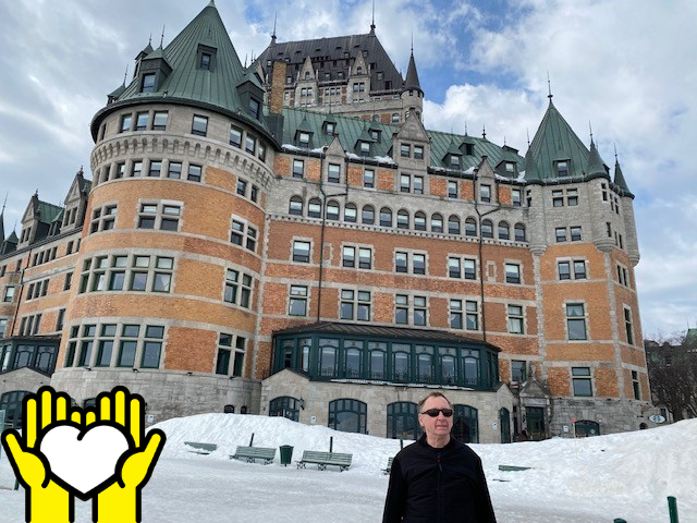 Une photo de Louis Lévesque debout en face du Château Frontenac à Québec. L'icône de deux mains tenant un cœur blanc sur un fond jaune, dans le coin inférieur gauche.