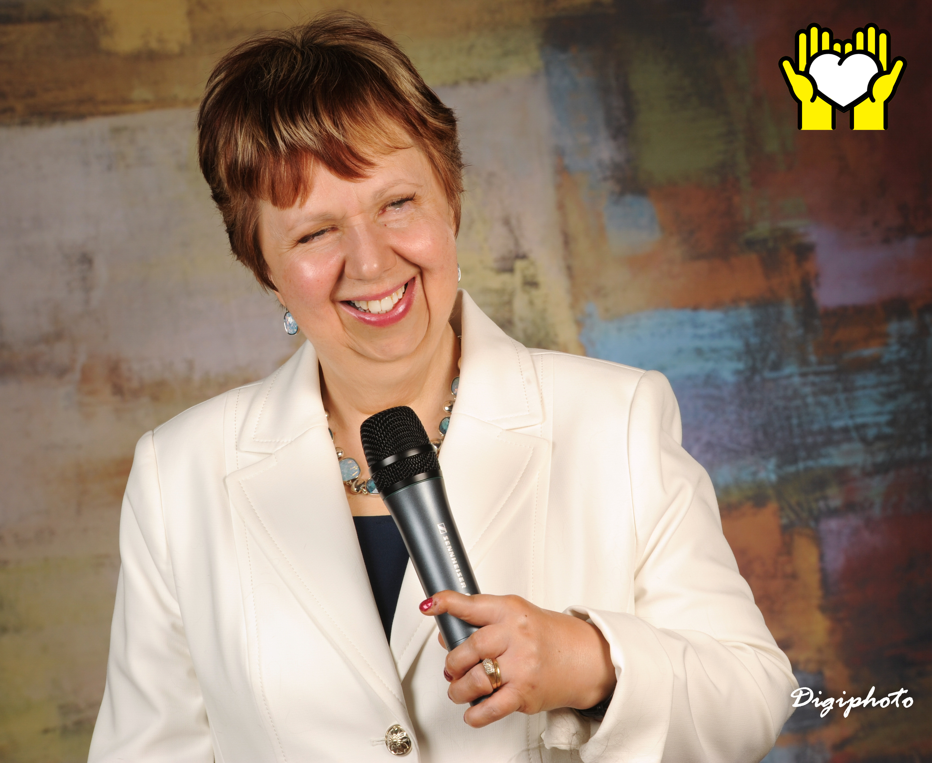 Une photo de Louise Burley souriant et tenant un micro de sa main gauche. L'icône de deux mains tenant un cœur blanc sur un fond jaune, dans le coin supérieur droit.