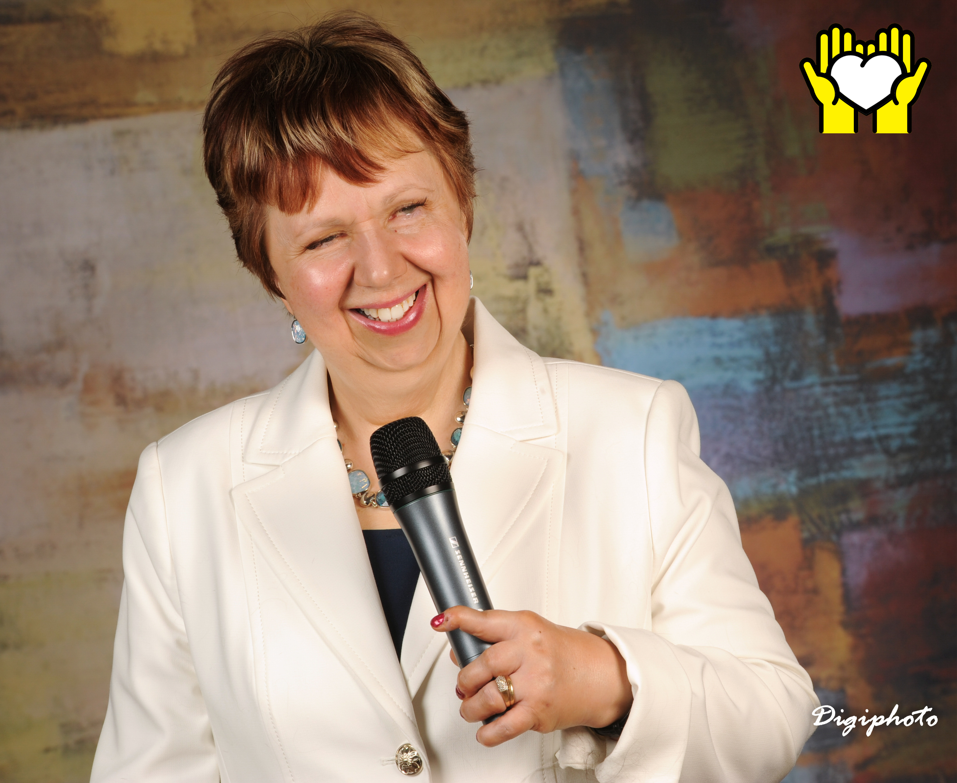 Une photo de Louise Burley souriant et tenant un micro de sa main gauche.  L'icône de deux mains tenant un cœur blanc sur un fond jaune, dans le coin supérieur droit.