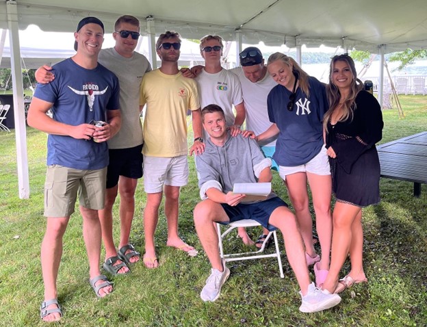 Five young men and two young women wearing shorts and t-shirts stand behind a young man (Matt) sitting in a lawn chair. They are outside on the grass under a tall white tent with the lake in the background