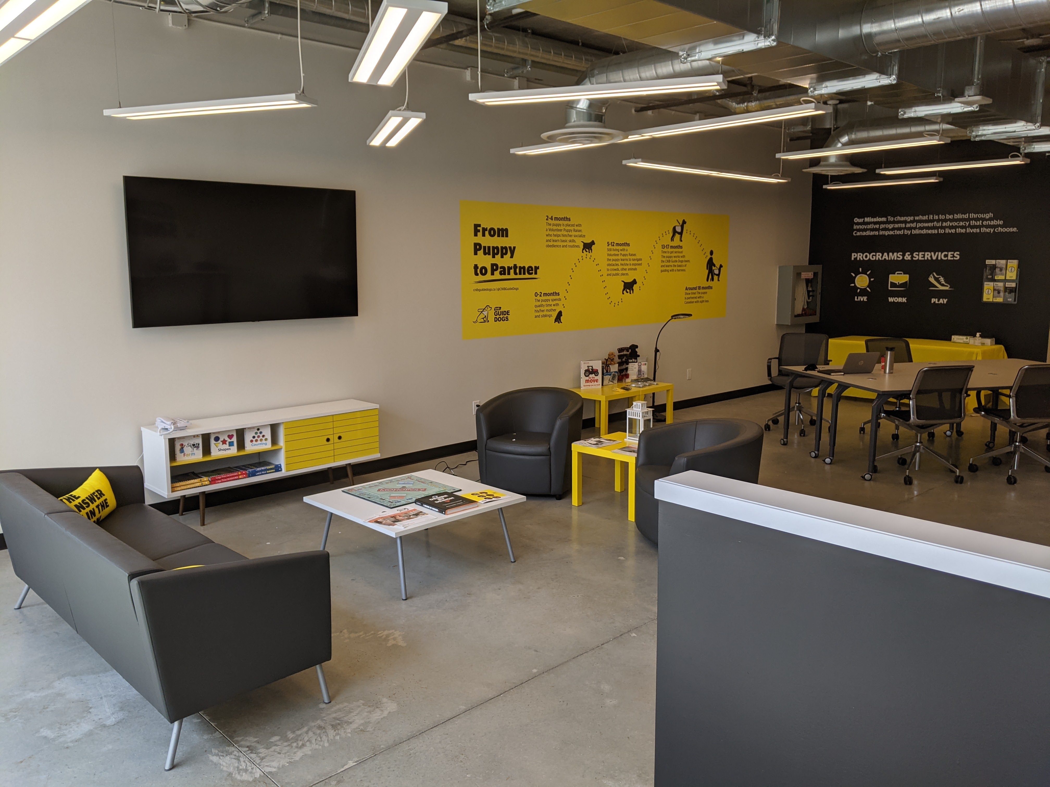 An image of the interior of the Barrie Community Hub. On the left, there is a sitting room with a couch, two chairs and a large television on the wall. In the  back of the room, there is a table and chairs where individuals might gather