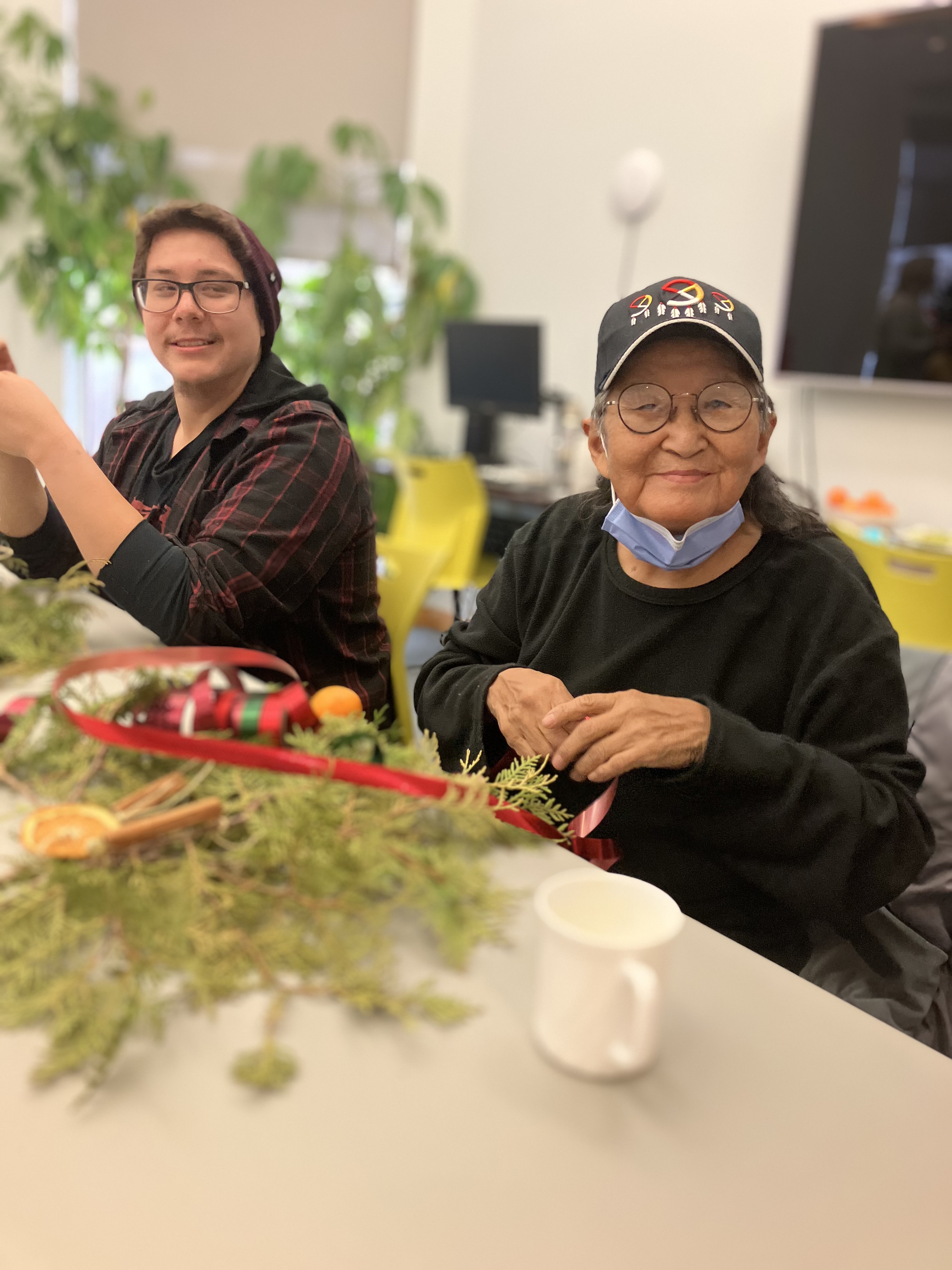 Deux participants autochtones souriants sont assis à une table au bureau d'INCA à Thunder Bay. Au cours d'un programme de vacances, elles créent des décorations avec des nœuds de cèdre frais, des tranches d'orange séchées, des biscuits de pain d'épice faits maison, des rubans, des cloches, des bâtons de cannelle et de la ficelle de jute.