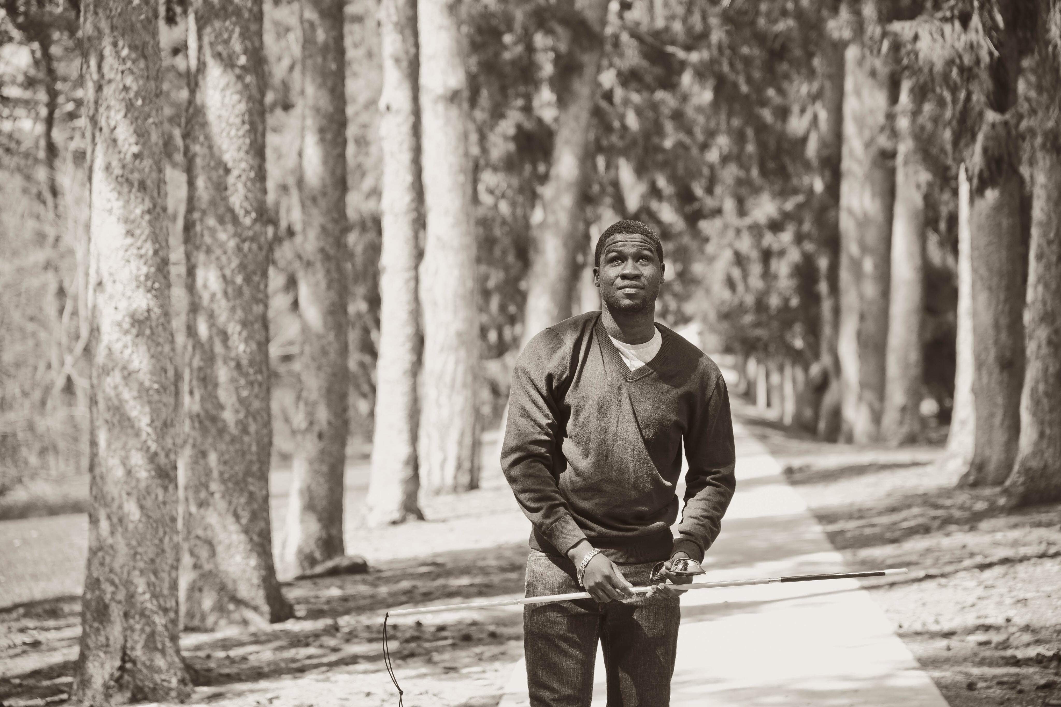 A black and white photograph of Ben walking in a forest. He holds his white cane up in the air near his waist.