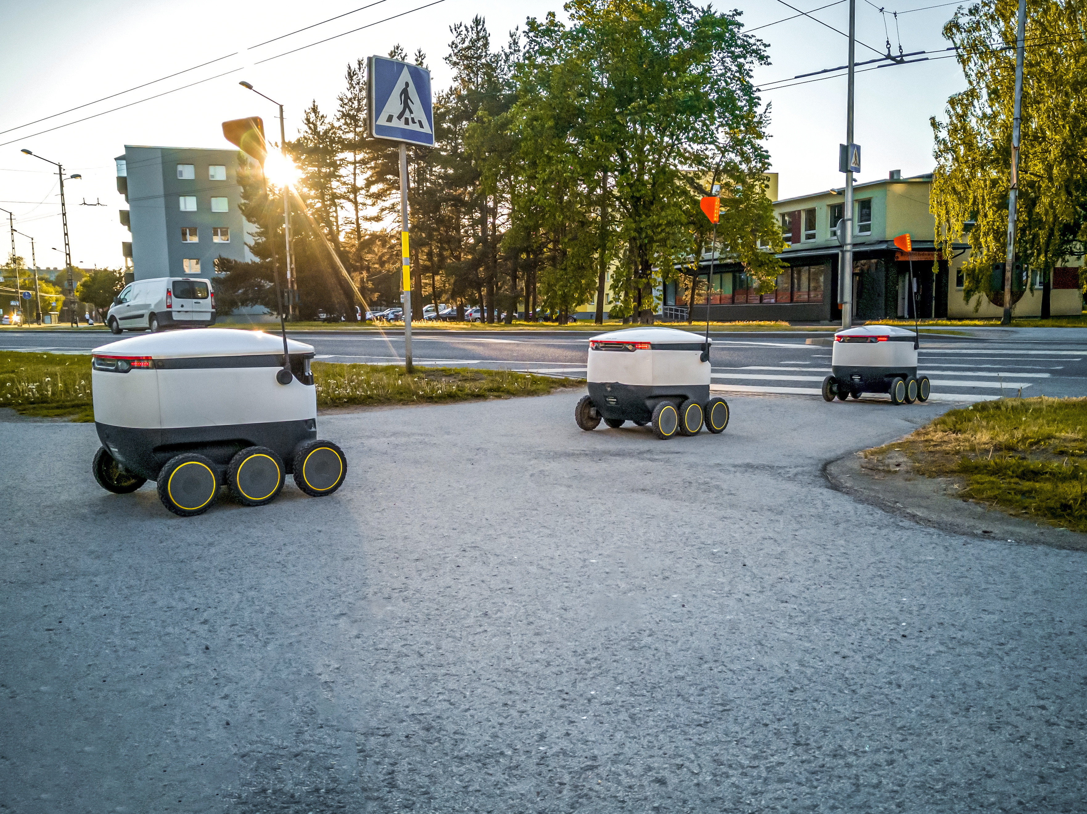 Three self-driving delivery robots drive along a city sidewalk.