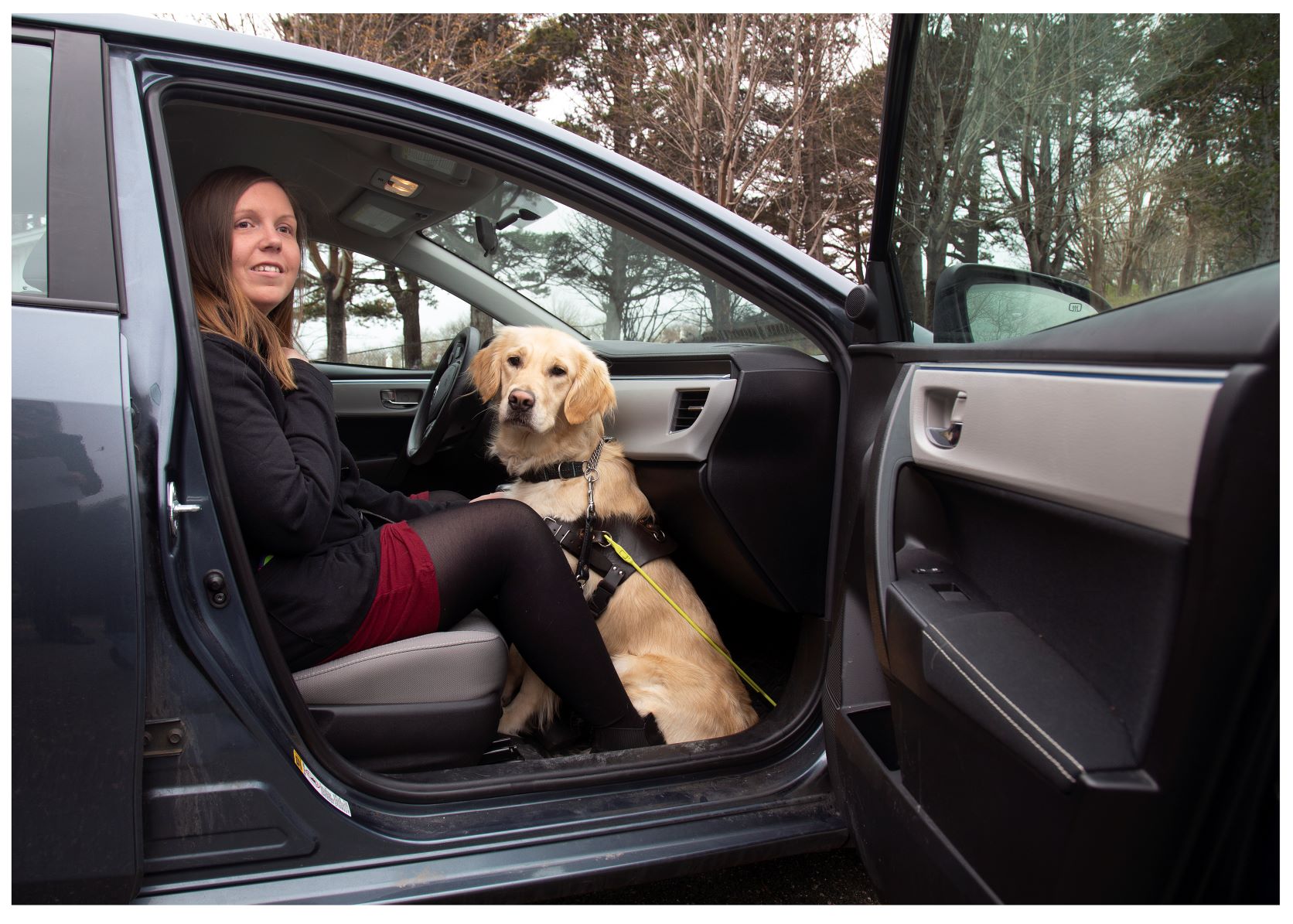 Une femme assise sur le siège passager avant d'une voiture dont la portière est ouverte; son chien-guide, un golden retriever, est assis à ses pieds.