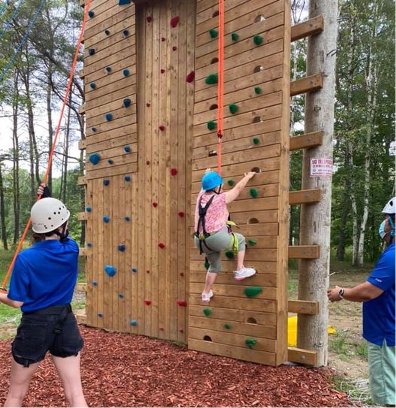 Fran Moreau in her helmet and climbing harness scaling the climbing tower while CNIB Lake Joe staff assist.