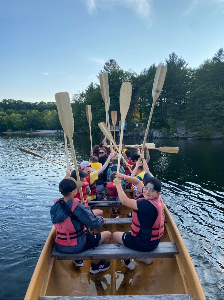  Du canotage au Centre Lake Joe! Des enfants et des jeunes sont assis dans le canoë Voyageur et tiennent leurs pagaies en l’air. 