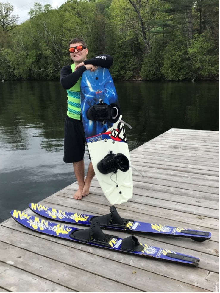 Josh est debout sur le bord du quai, vêtu d'une chemise et d'un short noirs, avec un gilet de sauvetage vert. Un ensemble de skis nautiques bleus et jaunes est posé sur le quai, et il est appuyé sur une planche nautique. Il sourit et porte des lunettes de soleil.