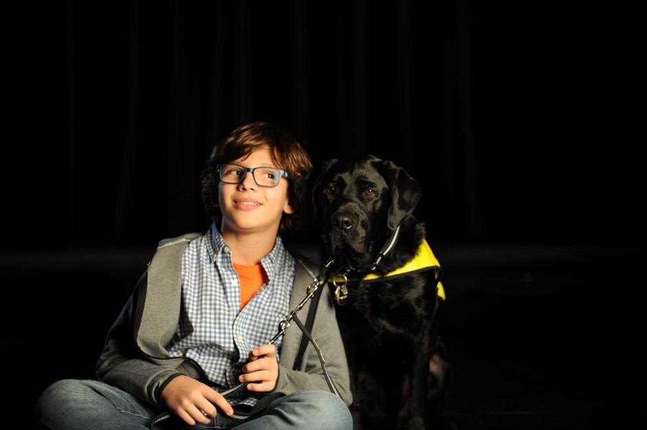 Gabriel sourit, assis à côté de Terry, son chien compagnon d'INCA, un Labrador retriever noir, sur le plateau de tournage du dernier message publicitaire de la campagne de collecte de fonds d'INCA.