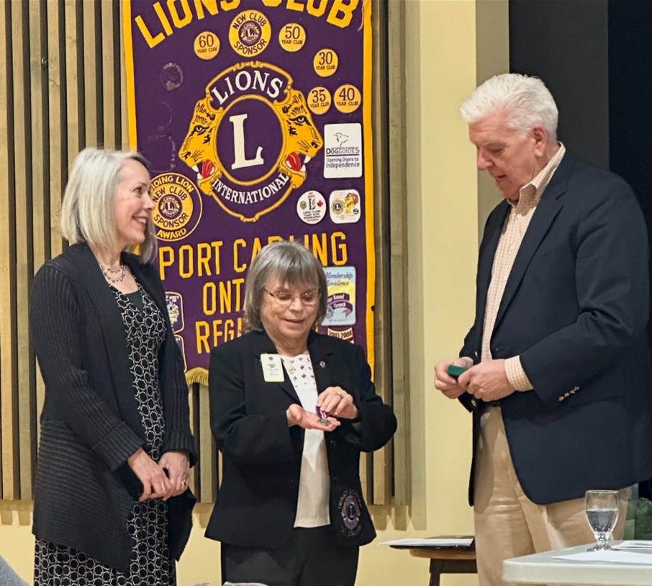 Sherri Helsdingen, Barb Ennis et le maire Peter Kelley devant une bannière du Lions Club de Port Carling. Barb tient la médaille dans sa main.