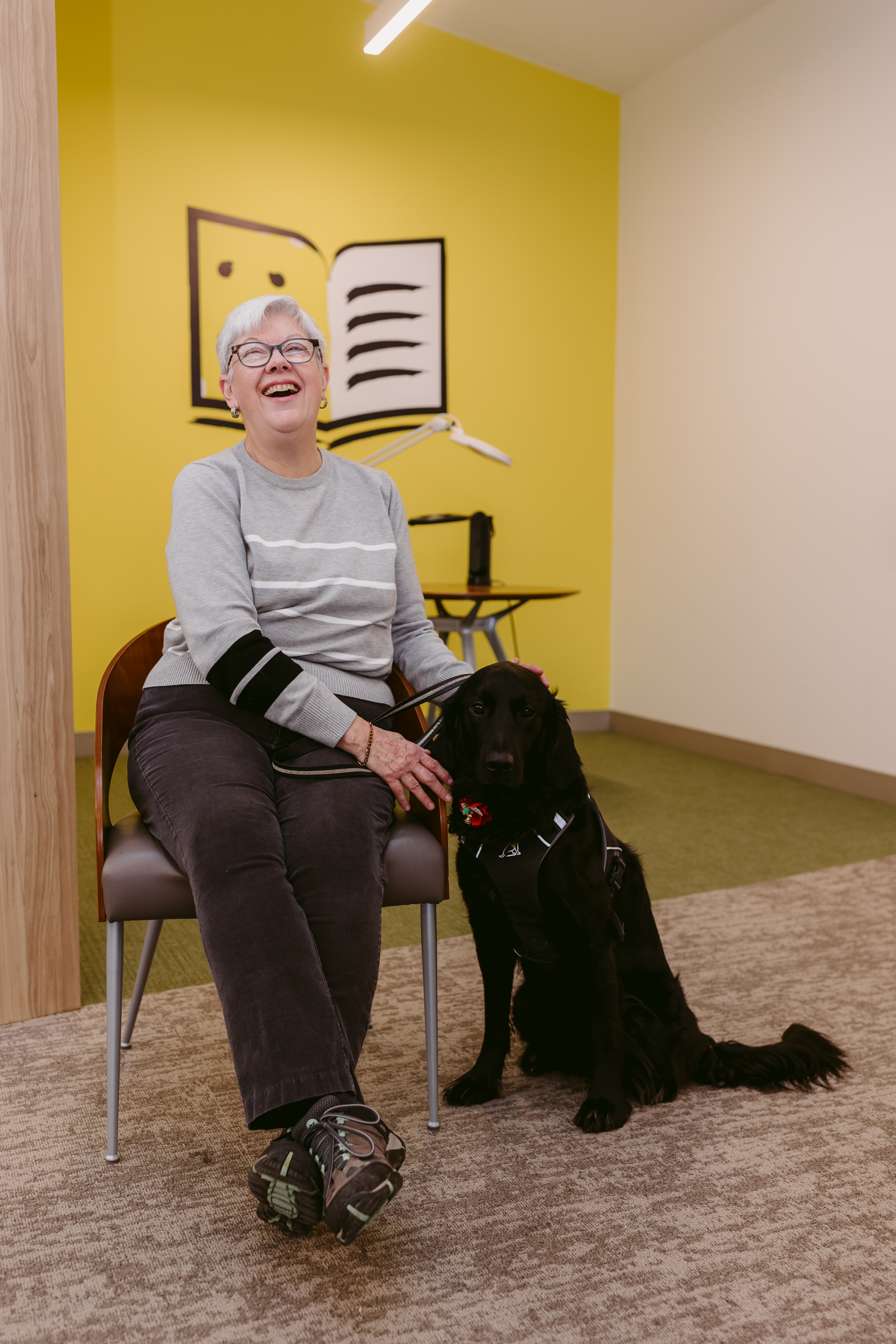 Dans un bureau d’INCA, Penny est assise sur une chaise. Son chien-guide, Honour, un croisement de labrador retriever noir et de golden retriever, est assis à ses pieds. Penny caresse la tête d’Honour et sourit de joie.