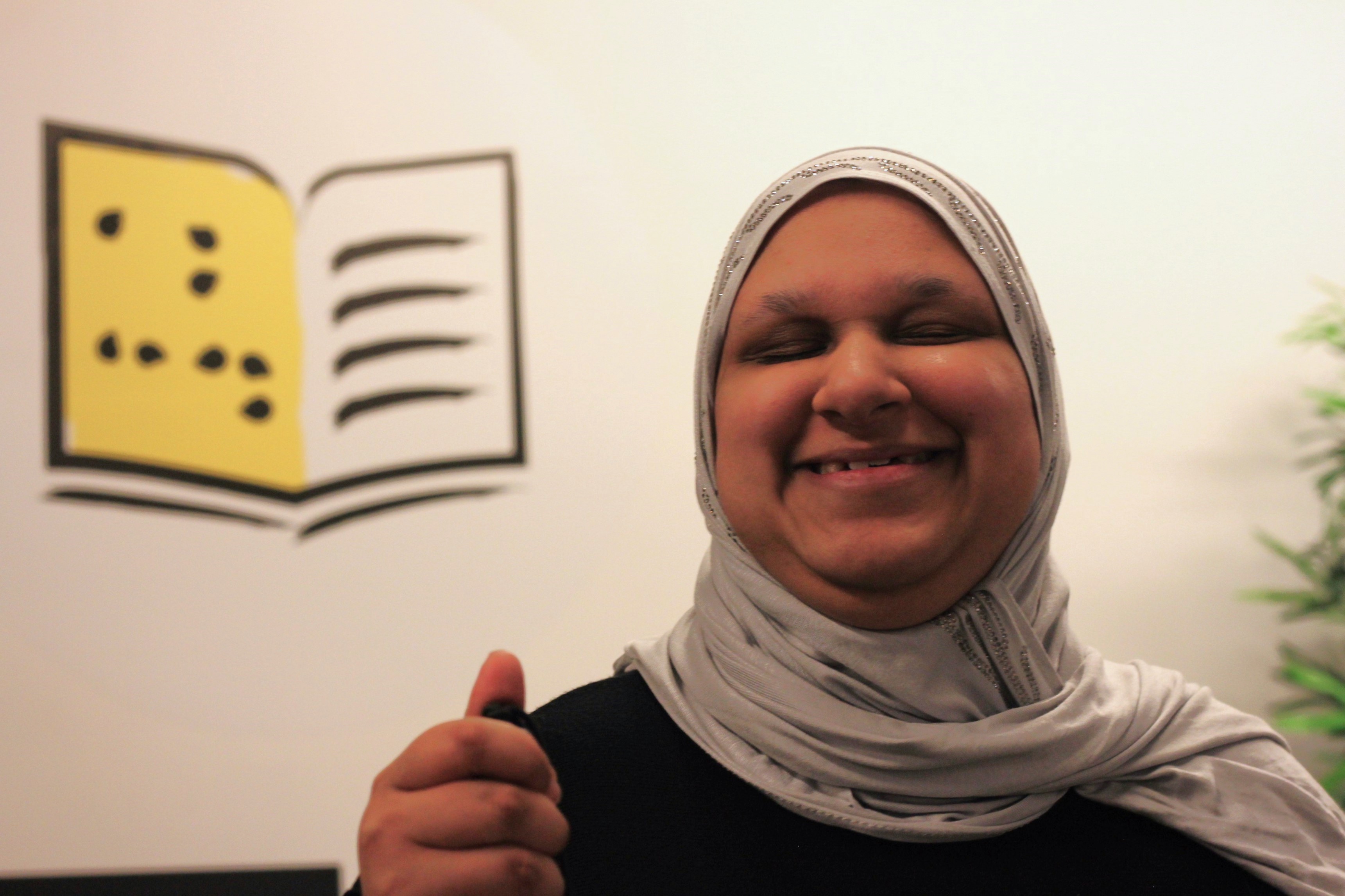 Salma tient sa canne en souriant. Derrière elle sur le mur l'icone des programmes Apprendre d'INCA )un livre en braille aux couleurs jaune et noir).