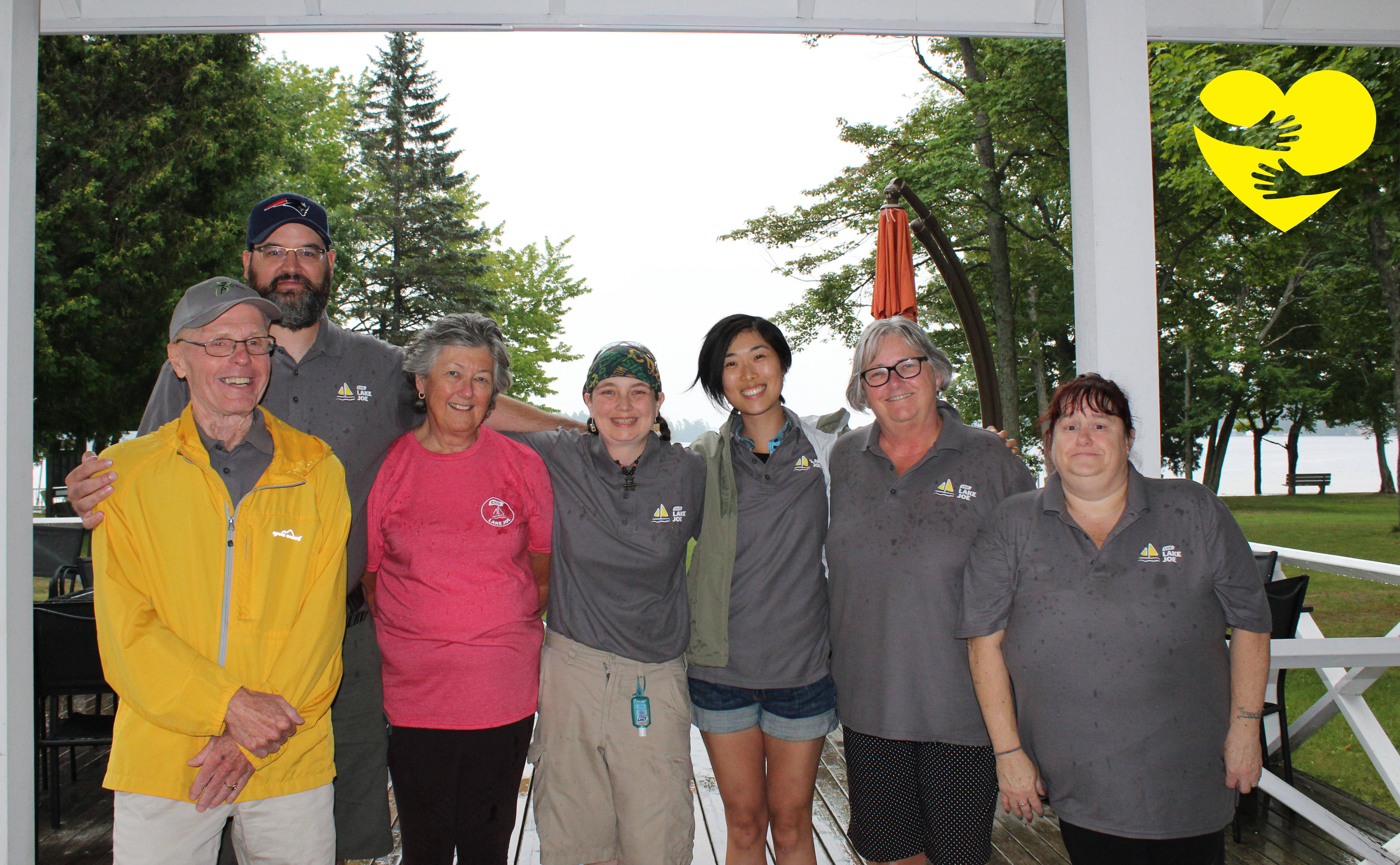 Sandra, portant une chemise rose, sourit pour la photo de groupe avec d'autres bénévoles d'INCA au camp Lake Joe. Un dessin de bras serrant un cœur jaune illustré se trouve dans le coin supérieur gauche de la photo.