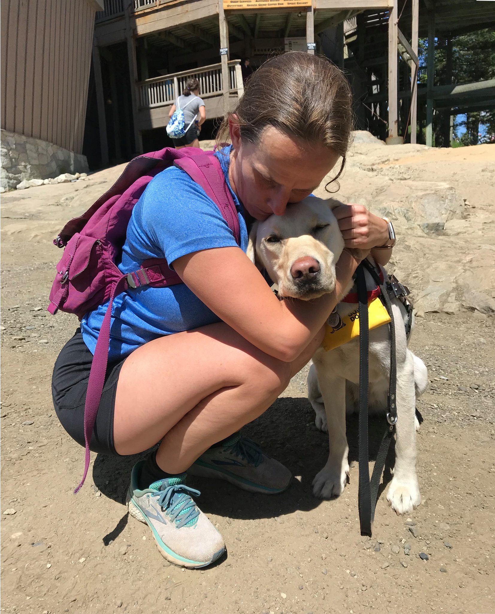 Sara, wearing hiking clothes, kneeling down giving Daisy a hug on a sunny day.