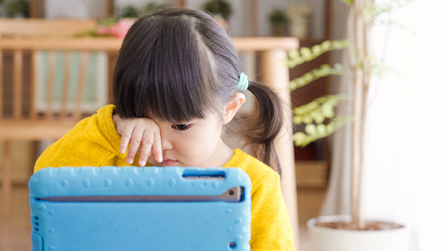 Une fillette aux cheveux foncés se frotte les yeux en regardant l'écran d'une tablette.