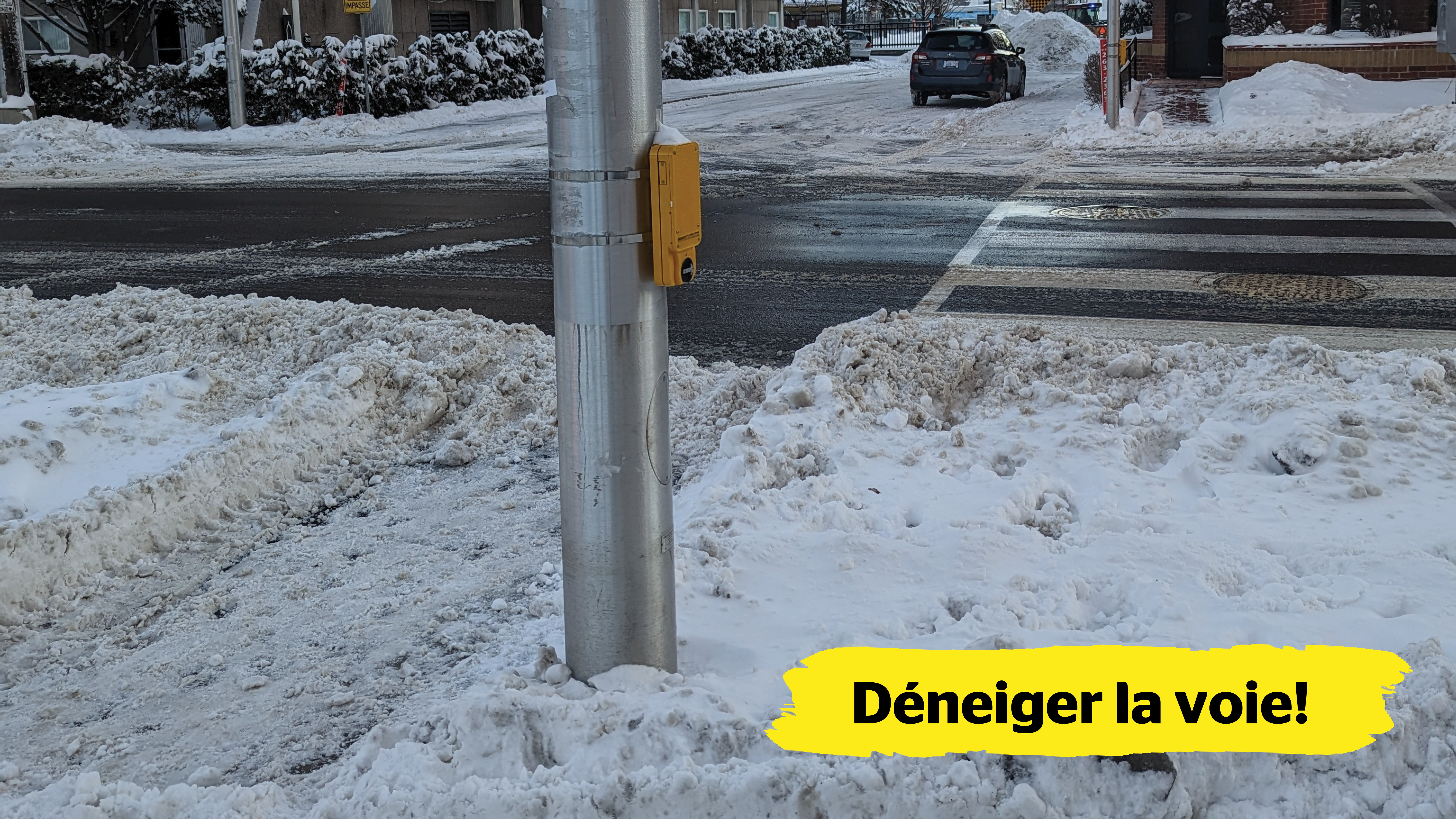 Un passage piéton est recouvert de neige et obstrue à la fois la voie de circulation et un poteau qui abrite le bouton-poussoir de la traverse de piétons.  Dans le coin inférieur droit de l’image, une bannière jaune affichant le texte « Déneiger la voie! » est superposée.