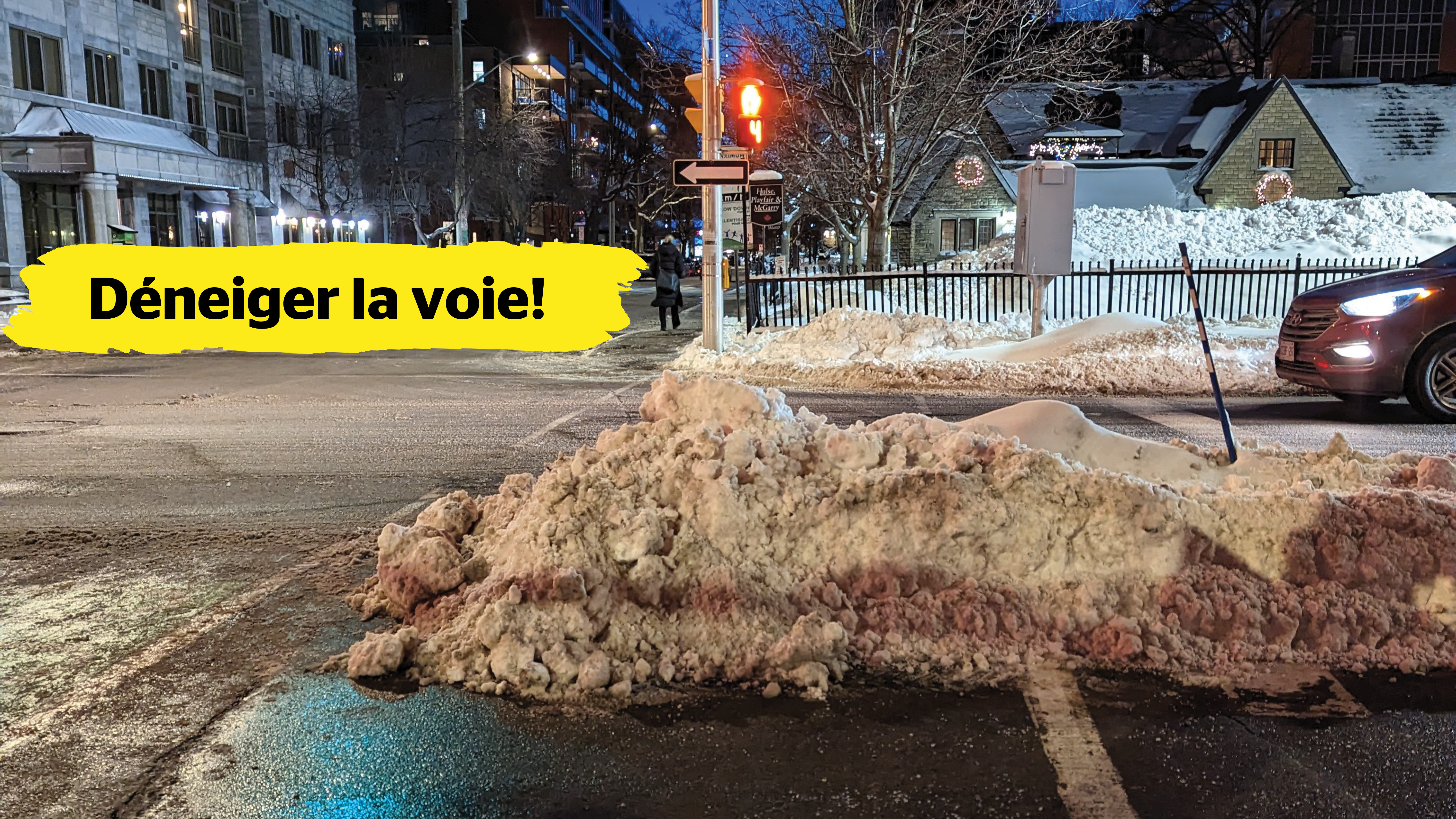 À des feux de circulation, un amoncellement de neige mal déblayé bloque l’accès à la moitié de la traverse de piétons. Dans le centre de l’image, une bannière jaune affichant le texte « Déneiger la voie! » est superposée.