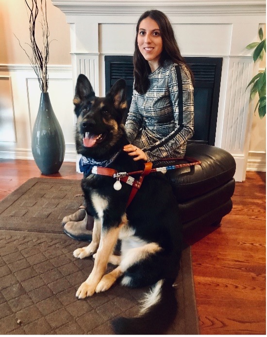 Stela sitting on a leather ottoman in front of a fireplace with her German Shepherd guide dog in front of her