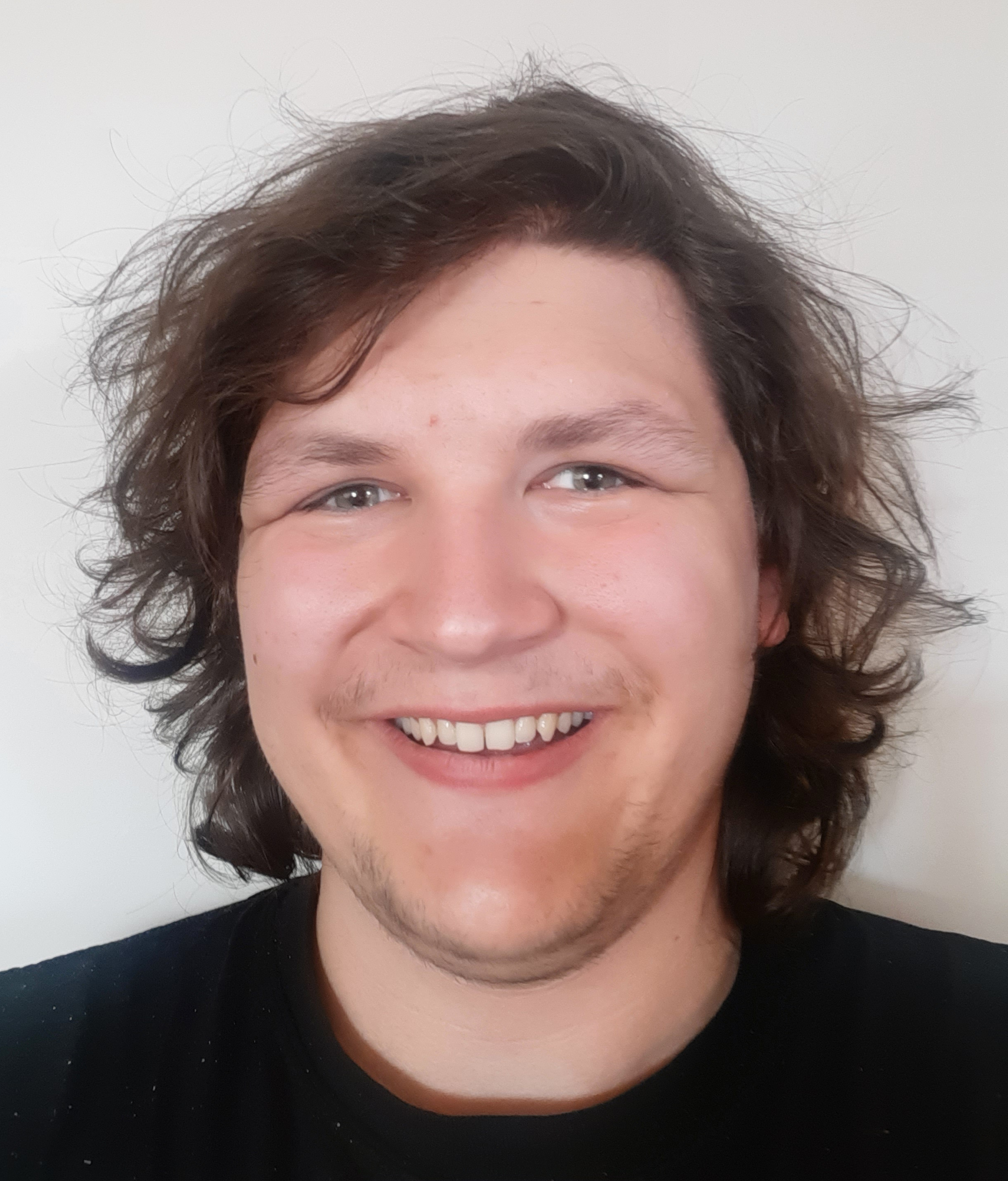 Tate, un jeune homme aux cheveux bruns, pose pour une photo de type selfie. Il porte un t-shirt noir et sourit.