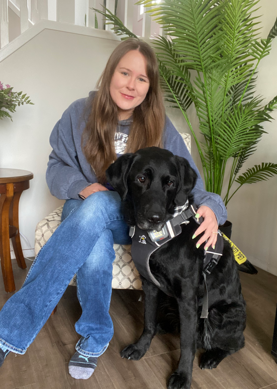 Taylor, une jeune femme aux longs cheveux bruns, est assise sur une chaise, le bras autour de son chien-guide Wallace, un Labrador noir qui porte un harnais et est assis à ses côtés.