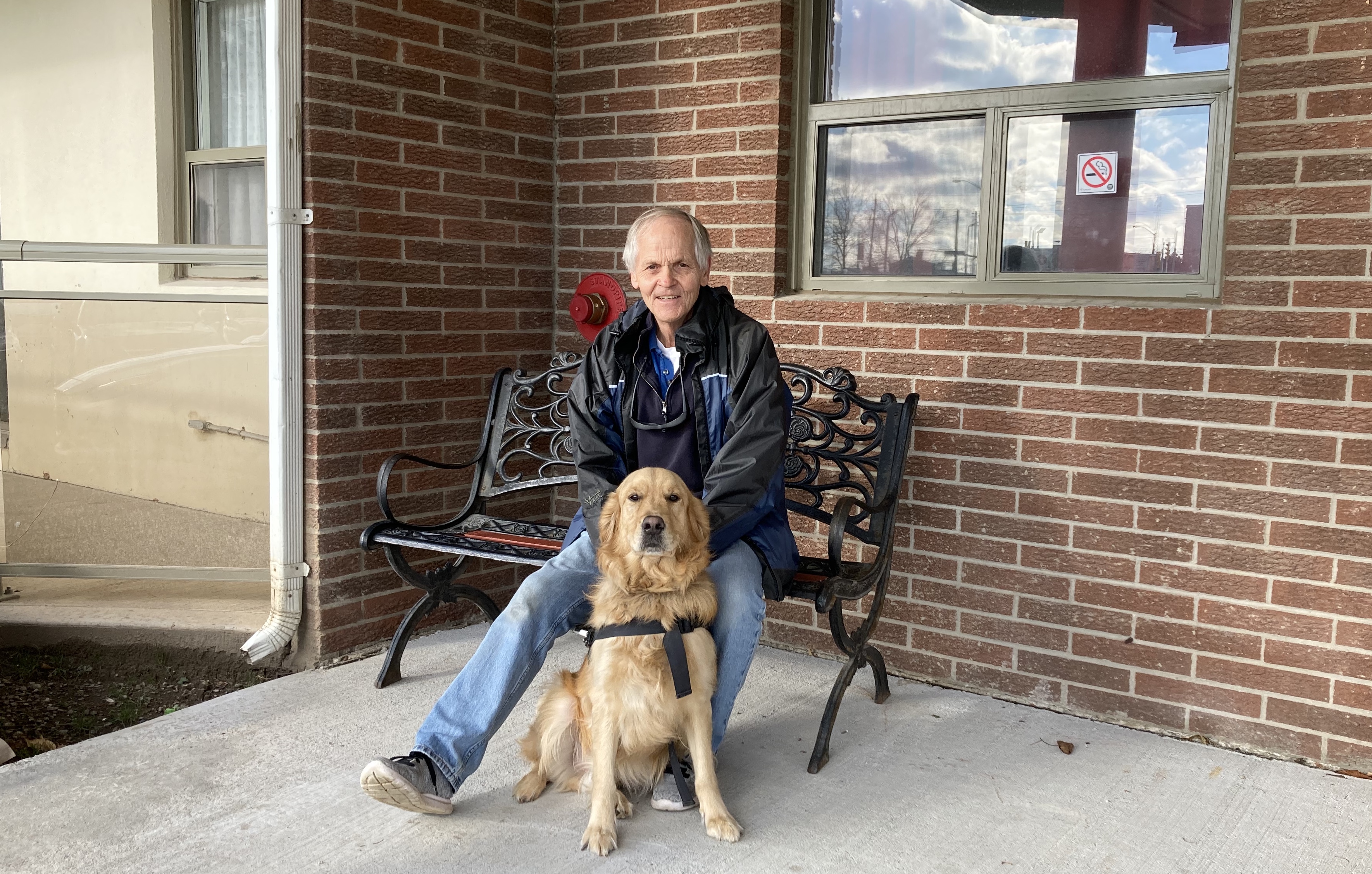 Photo of Terry sitting outside on a metal bench, smiling for the camera with Bert sitting front of him. 