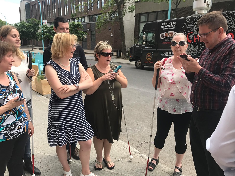 Groupe de personnes aveugles avec leur canne ou leur chien-guide, sur leur téléphone cellulaire sur le trottoir, souriant.