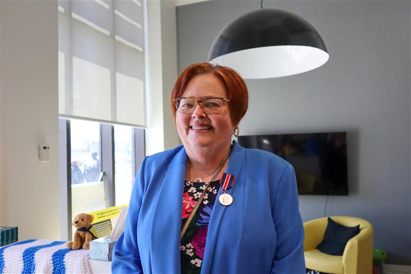 Cindy, a light-skinned woman with short red hair wearing a floral dress and a bright blue blazer, smiles at the camera.