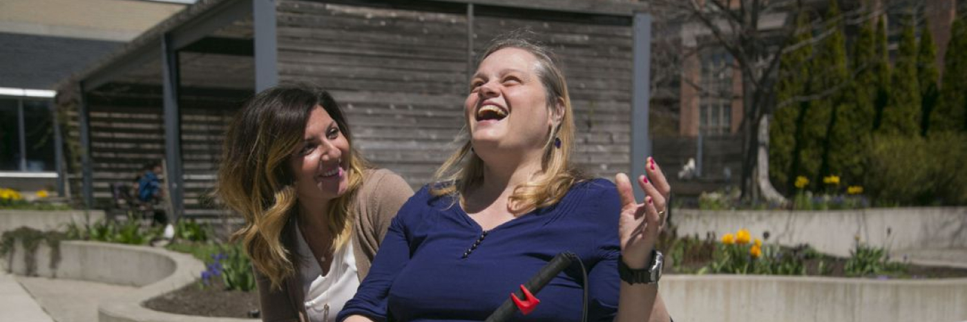 Une femme brune assise souriante et attentive aux côtés d'une femme blonde souriante tenant une canne blanche souriante. Elles sont toutes deux assises à côté d'un jardin olfactif. 
