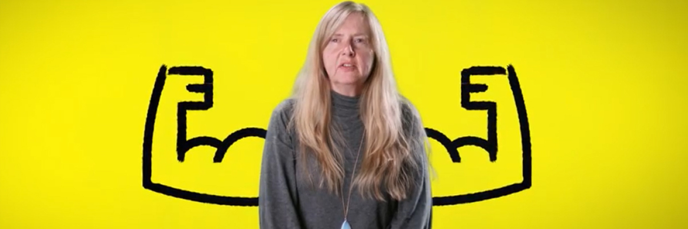 Diane Bergeron se trouve devant un arrière-fond jaune duquel se détache une illustration caricaturale de bras en flexion chaque côté d’elle.