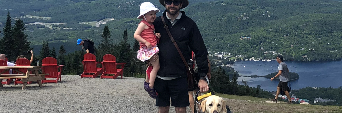 David avec sa fille dans un bras et de l’autre main, tenant dans son harnais son chien-guide Lilo, souriant pour la caméra, le Versant Sud du Mont Tremblant au Québec en arrière-plan