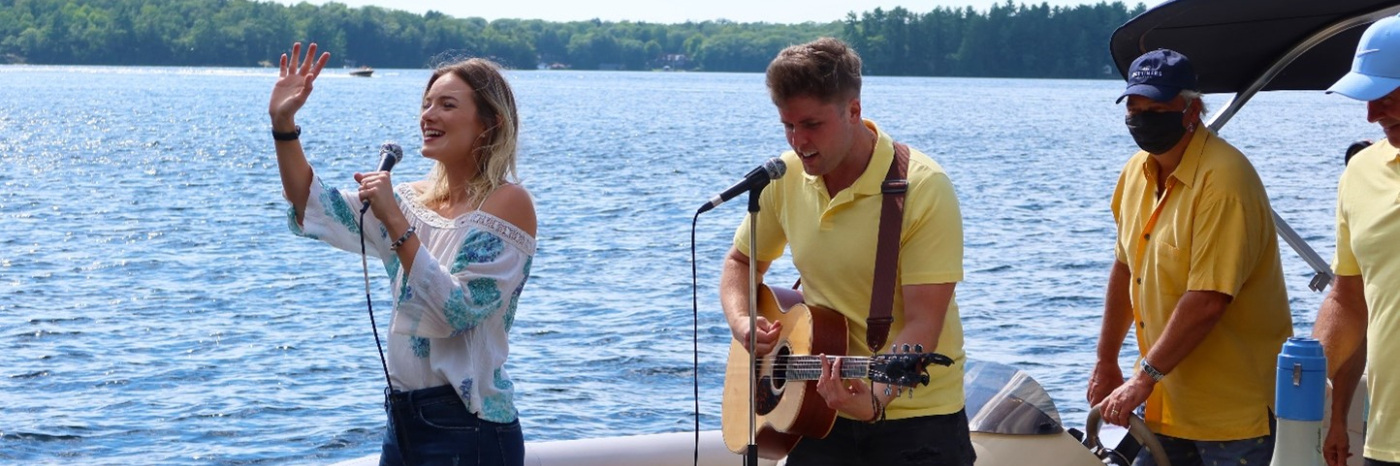 Leah Daniels chantant et saluant les spectateurs avec Will Hebbes à ses côtés jouant de la guitare sur un ponton scénique, divertissant les invités des quais sur le lac Muskoka. 