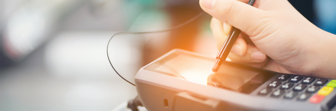A close up of a person’s hand using a stylus pen to make a selection on the touch screen surface of a payment terminal.