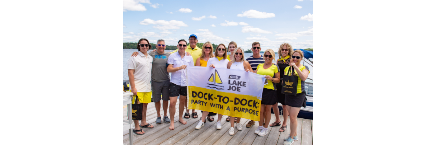 Des bénévoles de l’événement Muskoka de quai en quai d'INCA posent au hangar à bateaux du camp Lake Joe d’INCA, tenant des drapeaux à l’effigie de l’événement