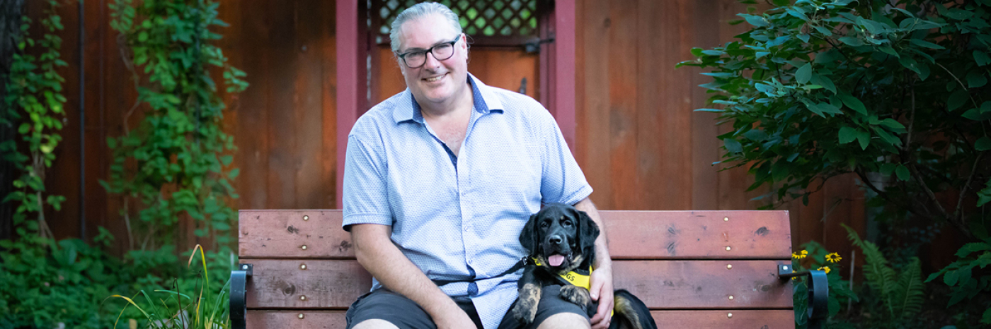 Christopher Adams assis sur un banc avec un très jeune Percy assis à côté de lui, portant sa veste jaune vif Future chien-guide d’INCA. 