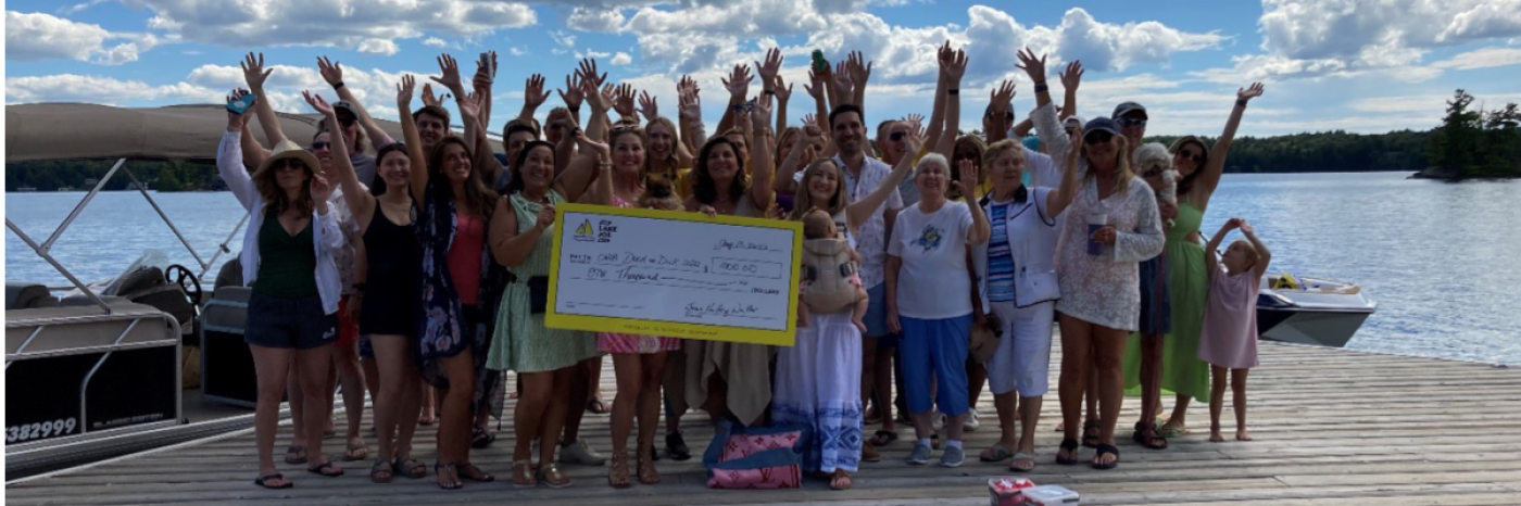 Un groupe d'invités debout sur un quai au bord de l'eau et salue avec enthousiasme, en montrant leur don d’un chèque géant de 1 000 $. 