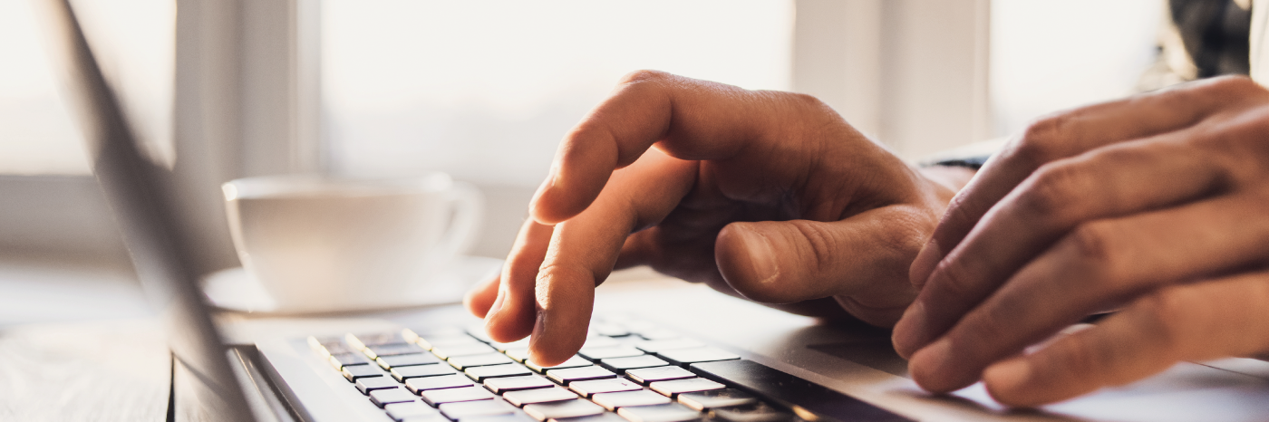 Gros plan sur les mains d'une personne tapant sur le clavier d'un ordinateur portable. L'accent est mis sur les mains et le clavier, l'arrière-plan est flou mais il s'agit d'un intérieur bien éclairé